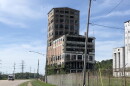 An abandoned, multi-story industrial building with broken windows stands behind a chain-link fence next to a highway.