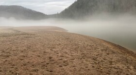 A barren muddle landscape shrouded in fog. Beneath the fog, barely visible, is Gatewood lake. In the background is forest.