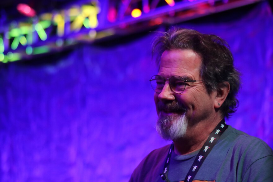 Phil Maass is shown smiling in front of a blue background, wearing a gray t-shirt and an IPR lanyard around his neck.