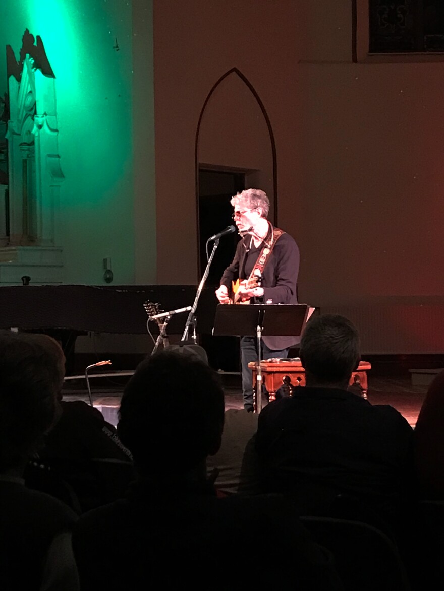 A man on stage in a former church singing and playing guitar. There is a green light shining on the upper left corner of the photo.