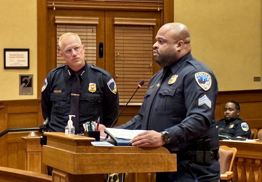A police officer stands at a podium speaking, while another officer stands behind him and a third officer sits at a desk in the background.