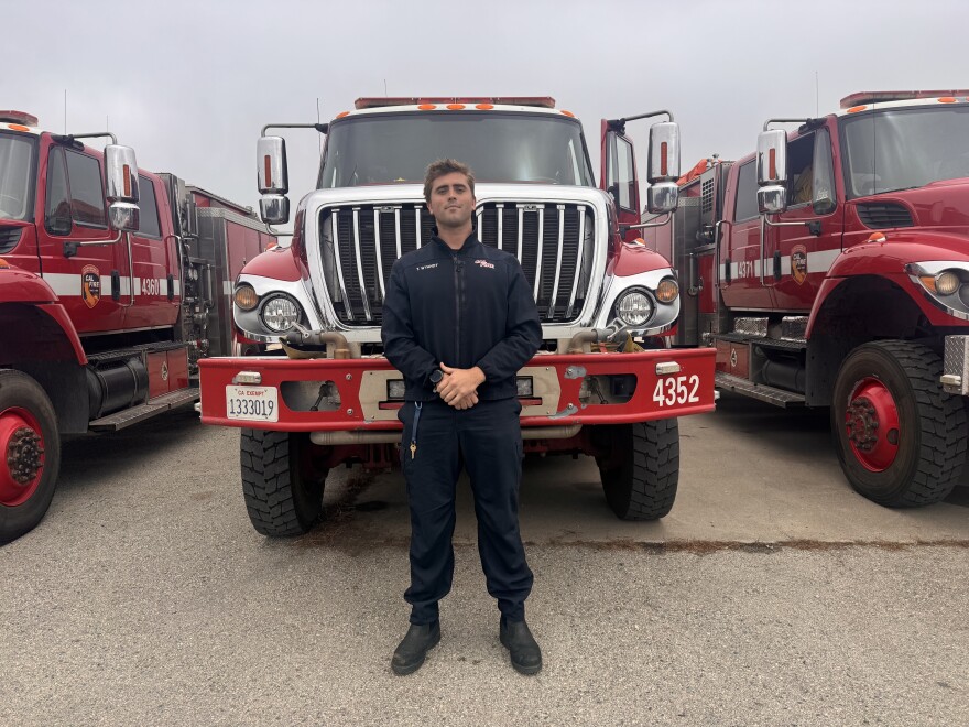 A man stands with his hands clasped in front of a row of fire trucks.