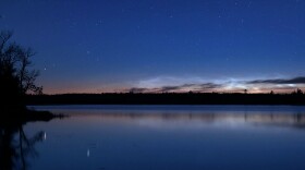 Noctilucent clouds on a tree-lined horizon beyond a lake and under a starry sky