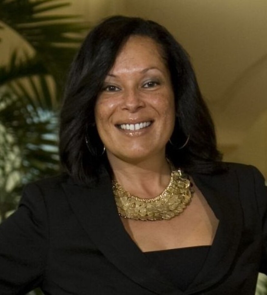 A headshot of a a woman with long dark hair smiling at the camera and wearing a thick, yellow necklace with several pendants dangling off of it.