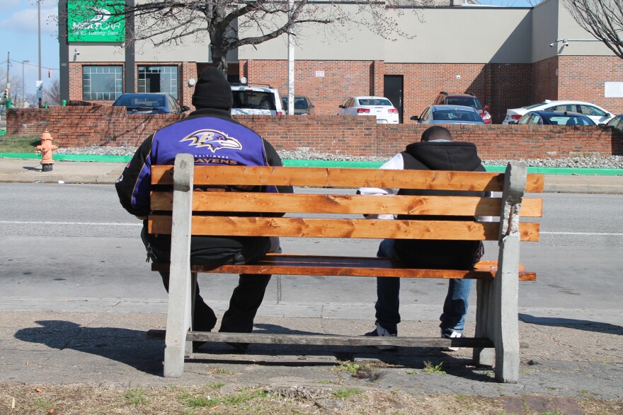 Two people wait at an unsheltered bus stop on North Avenue. City education advocates say that long MTA commutes are a barrier for Baltimore students.