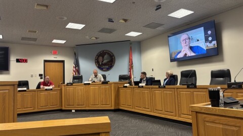 Four people sit at a daias in the Jefferson City Municipal Building meeting room. Three men are to the left, and one woman is to the right. A council member's face appears on a screen behind the woman; he and few others are joining the meeting on Zoom.