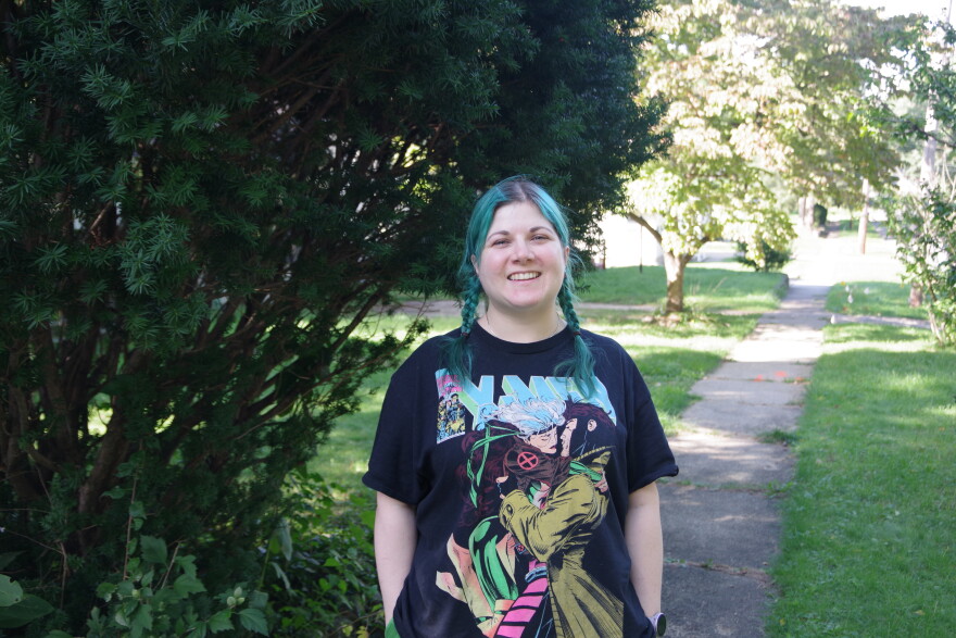 A woman with two blue braids stands on a sidewalk in front of a large bush. She's wearing a black t-shirt that shows two people embracing each other for a kiss. "X-Men" is written at the top of the shirt. 