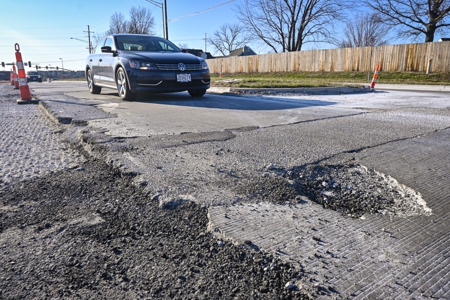 Grooved, mottled pavement is seen in the foreground of a four-lane road. There is a large pothole in one lane of traffic where a car is about to travel. Orange pylons can be seen nearby marking off the road that has been stripped for resurfacing.