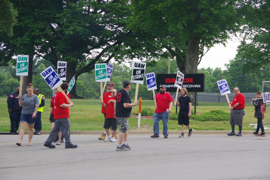 Around ten people stand on the side of the road in front of a chain-link fence and a black sign to the middle right of the image that reads "DAUCH" in red text, below which reads "Shipping & Receiving" in white text with an arrow pointing to the right. The men and women hold different colored signs, but most of them read the same message; "UAW ON STRIKE." One woman's sign to the far right of the image can only be partially read, it says reads "UNFAIR LABOR PRACTICE" in blue text.