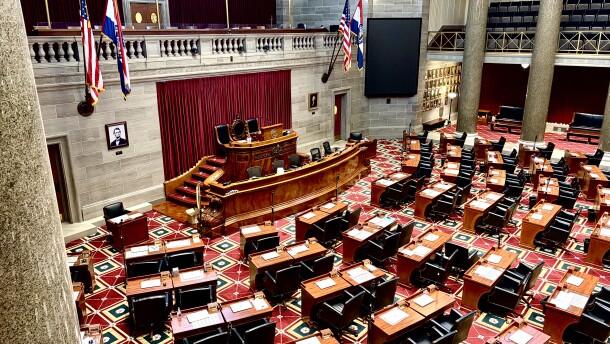 A view from the gallery of the Missouri House of Representatives floor. Empty wooden desks and dark chairs face the dais. 