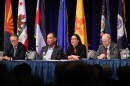Three women and a man sit on a stage in front of state flags