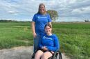 A woman standing and a teenage girl sitting in a wheelchair in the middle of a field. Both wearing a blue shirt.