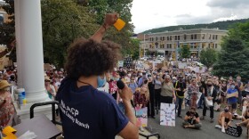 A person wearing a mask holds up a fist while speaking into a mic in front of a crowd of hundreds on a lawn below some steps