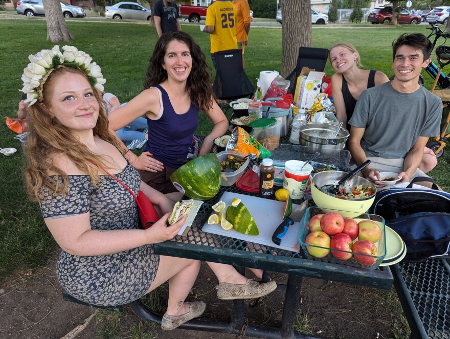 A group of young adults smile and pose for a photo sitting around a picnic table at a park. The table is filled with fruit, salads, and other homemade dishes.