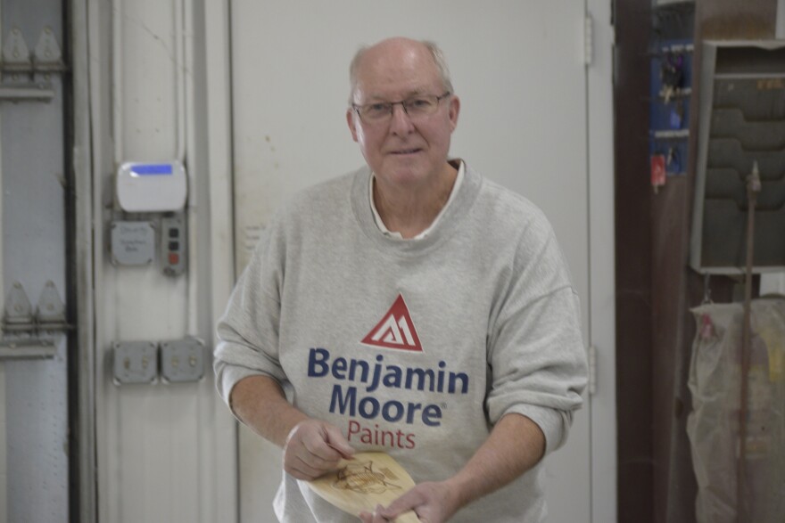 An older man wearing glasses and a "Benjamin Moore Paints" sweatshirt holds a wooden item in a workshop, standing near a white door and some tools.