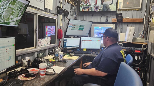 A man in a blue shirt and ballcap sits surrounded by computer screens. 