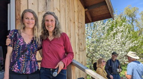 Two women stand on the stairs of a wooden building with an apple tree and other people in the background.