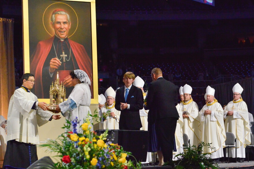 A family present a relic to a priest at a ceremony with a large portrait of the late Archbishop Fulton Sheen in the background and a gathering of Catholic bishops behind them.
