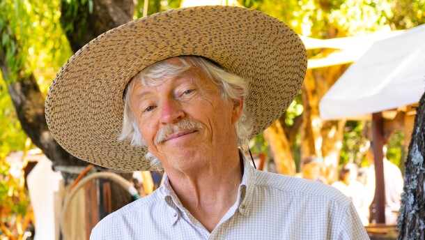 A man in a checked shirt and a wide brimmed hat poses in front of a vibrant outdoor background.