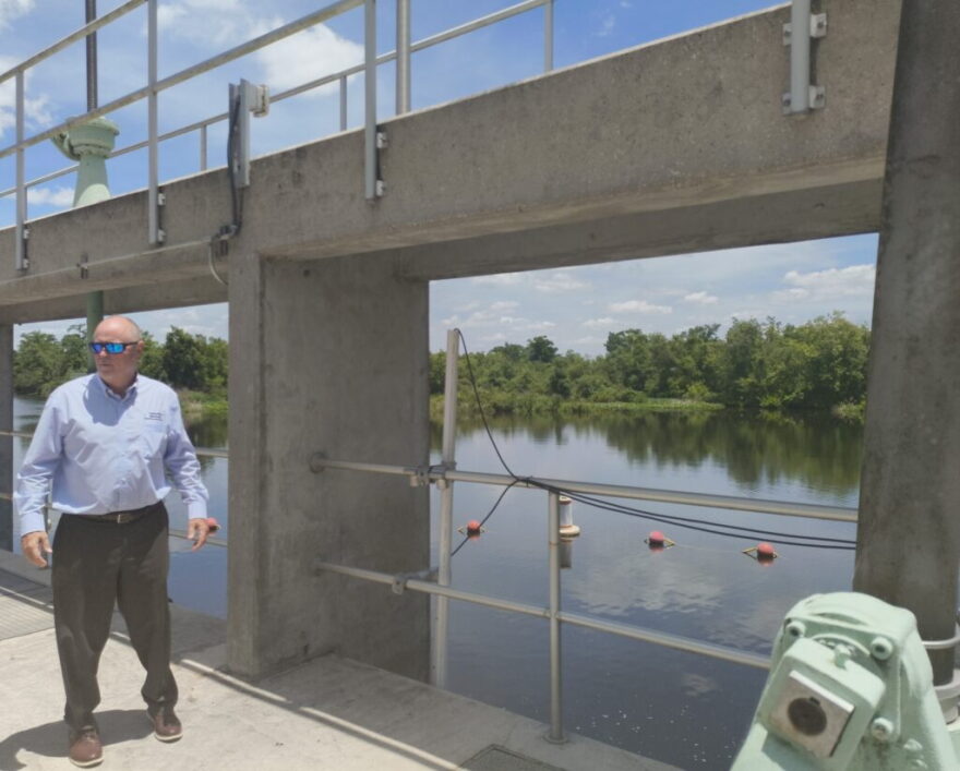 man in blue shirt and dark pants stands on a walking area over a large body of blue water, another higher walking area is next to him