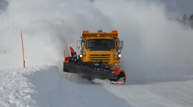 A yellow snow plow moves snow along a stretch of roadway. 