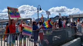 Supporters wave signs on a bridge on a sunny day. Many are wearing tie dyed shirts and rainbow colors.