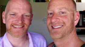 A photo of two brothers smiling. One is wearing a purple button down shirt. The other is wearing blue and black tank top