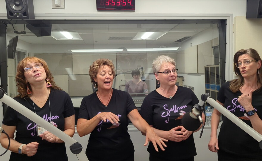 Four women wearing shirts that say "saffron" singing inside a radio studio. 