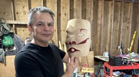 Micah McCarty stands in his workshop wearing a black T-shirt and holding up a nearly-finished mask that is carved from cedar and painted with red lips and eyebrows. It has abalone-inlaid white eyes. He is grinning an proudly displaying his ar. The workshop is full of sawdust and jumbled tools are everywhere.