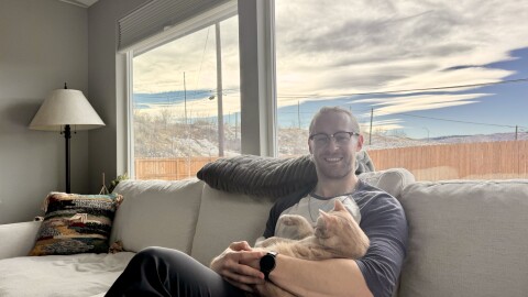 A man sits holding a cat on a grey living room couch in front of a window with a blue sky.