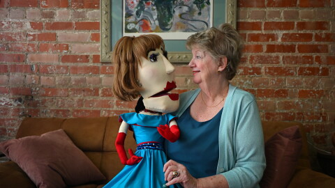 A woman sits on a couch next to a brightly colored puppet