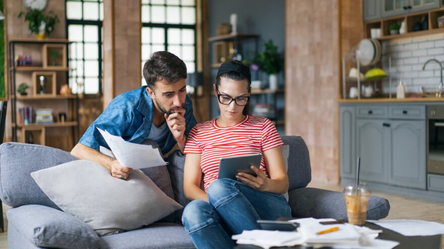 Couple calculating bills at home using laptop and calculator. Young couple working on computer while calculating finances sitting on couch. Young man with wife at home analyzing their finance with documents.