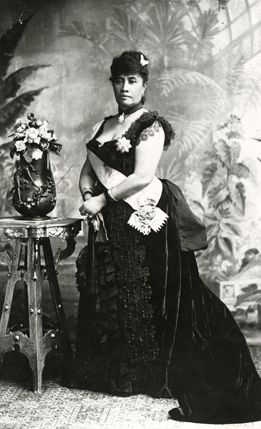 A black and white photo of Queen Lili'uokalani. She stands in front of a background of foliage in a beautiful velvet gown with a white sash.