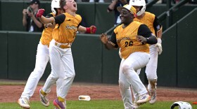 Four kids in baseball uniforms jumping up and smiling after winning a game on a field.