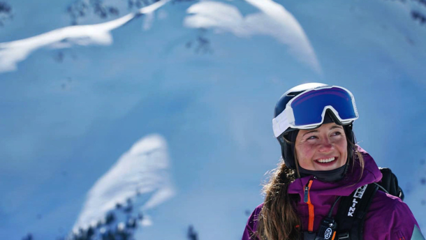 A woman wearing a ski helmet, goggles, jacket and backpack smiles. Behind her is a massive snowy peak dazzled by sunlight.