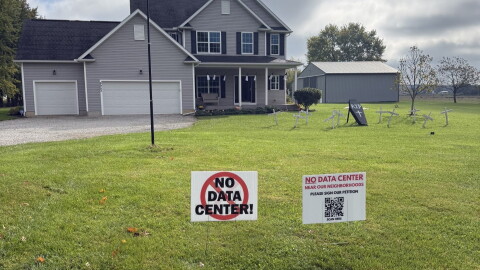Two signs in front of a large residential home on a rural plot read "No Data Center!" and "No data center near our neighborhoods, please sign our petition," with a QR code.