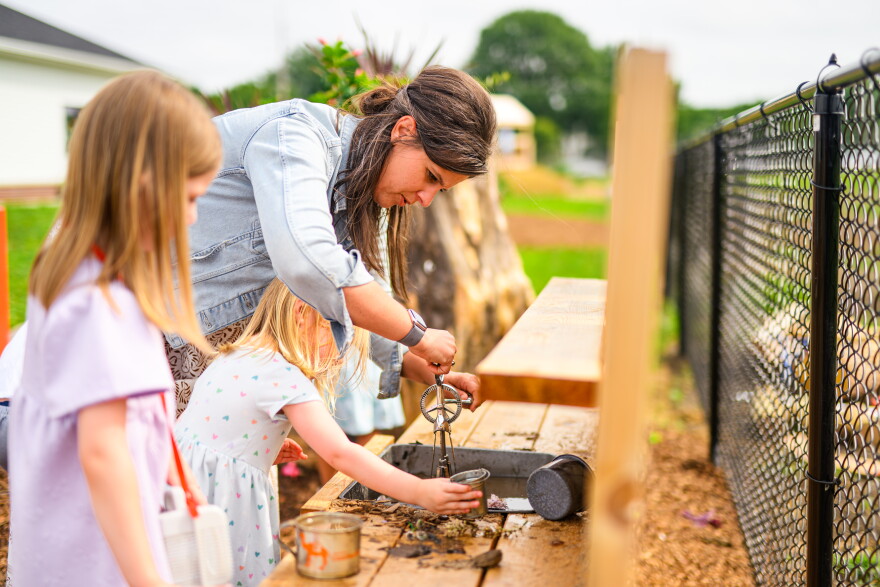 A woman and two young girls play and explore with water and metal tools on a wooden table outdoors near a black chain-link fence.