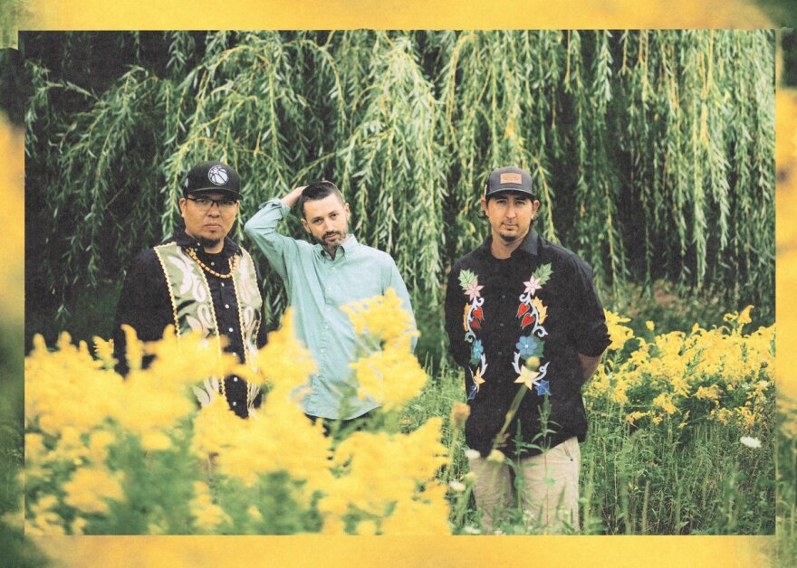 Three men standing in a field of yellow flowers with a willow tree behind them.