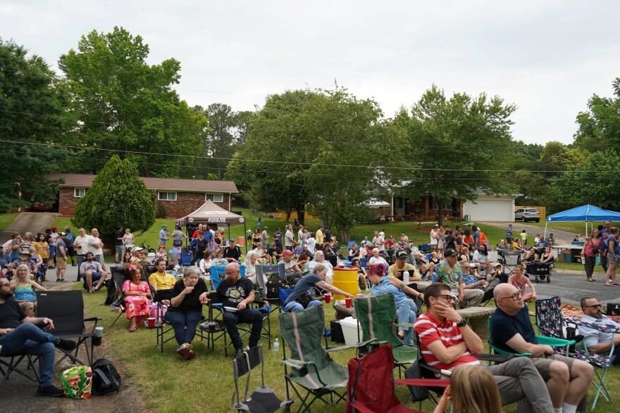 People gather outdoors in lawn chairs and around tents during a neighborhood festival.