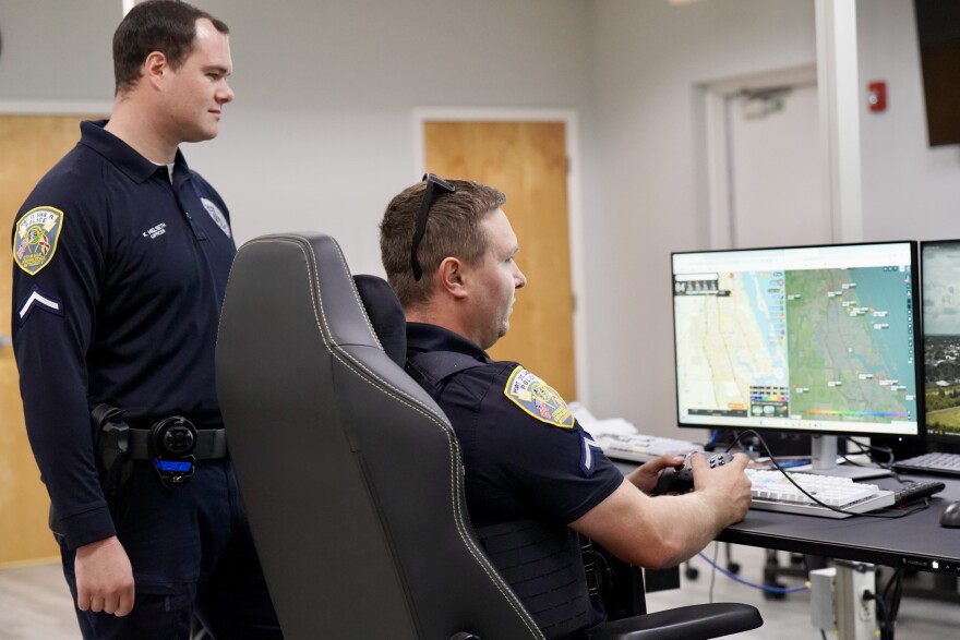 PSLPD officers in uniform operate drone equipment remotely from behind a computer screen at the station. One officer sits in a chair holding a controller with maps on the screen, the other stands behind his chair looking at the screen
