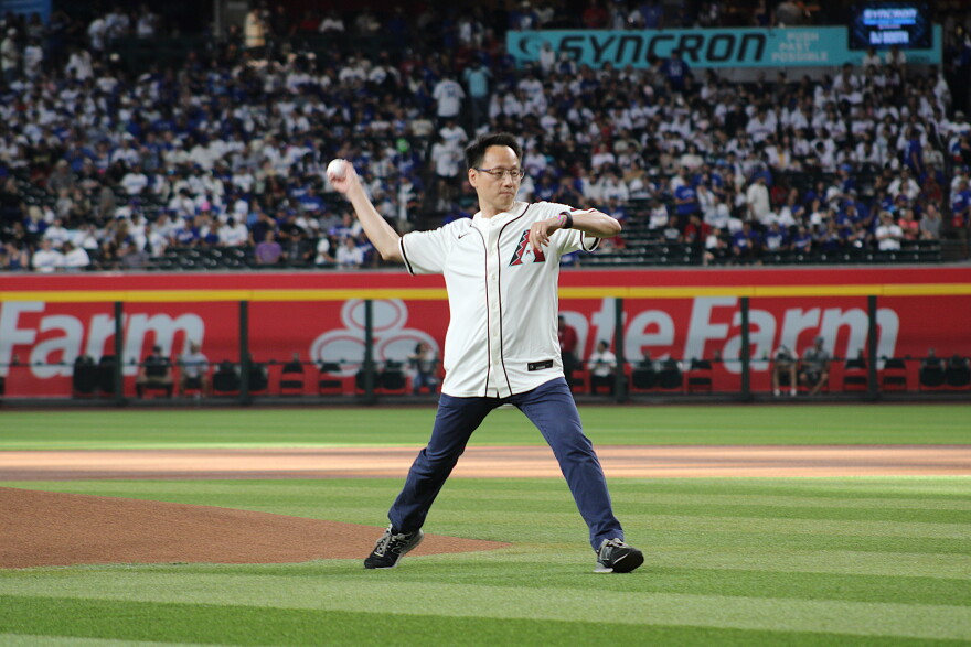 A man in an Arizona Diamondbacks jersey and jeans throws a baseball pitch in the middle of a baseball field. Fans sit in the stadium seats behind him in the distance.