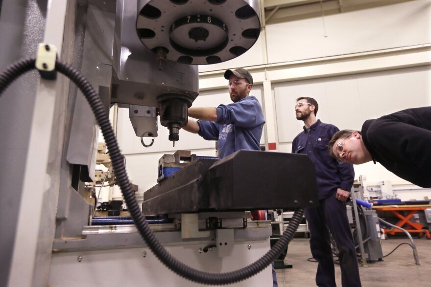 At the Lansing Community College, Brad Bancroft, wears a faded blue hoody and a camo baseball cap. His sleeves are rolled up as he operates a mill. Another man stands behind him in a deep blue button up and matching pants, watching Brad as he operates the machine. Another person leans into the shot from the right, he wears a black hoodie with the same safety glasses the others wear. He appears to be looking closely as the drill moves towards the object being machined. 