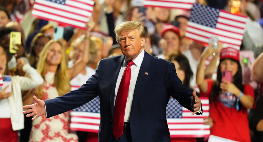 President Donald Trump stands with his arms outstretched. A crowd behind him waves American flags.
