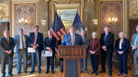 A man in a gray suit stands at a podium in front of a few other people, an American flag and the State of Utah flag