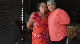 Two women pose for a portrait. They are hugging at the shoulders inside a darkened house.