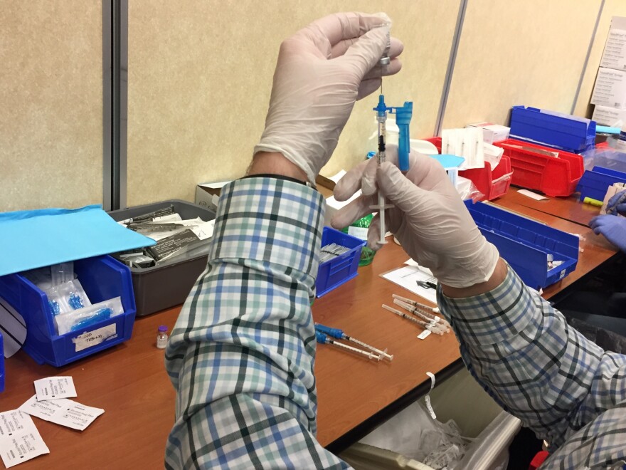 A person wearing a plaid long sleeve shirt and white gloves fills a syringe with Pfizer's COVID-19 vaccine over a table laden with alcohol swabs, more syringes and other supplies.