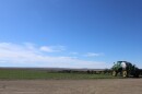 Wide, open landscape with a large agricultural sprayer parked in a grassy field under a vast blue sky. The sprayer has extended horizontal booms spanning much of the frame. A dirt road runs across the foreground, and low hills and sparse structures appear in the distance, emphasizing the rural setting.