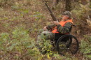 A hunter using a folding-frame wheelchair sits in a wooded area during fall. He is holding a shotgun and wearing camouflage clothing with a bright orange safety vest and camouflage cap.