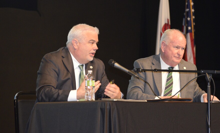 Two men in suits sit at a table on a stage with a microphone between them.