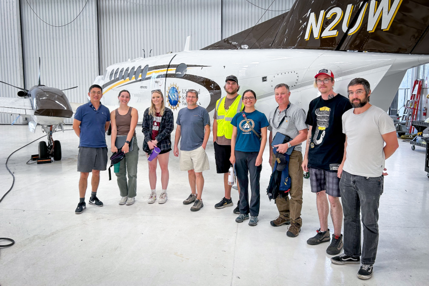 A group of nine people stand in front of a small aircraft in a hangar.